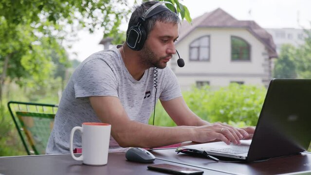 A man in headphones works on a laptop outdoors in the garden. Male programmer communicates online via video link. Distant work. 