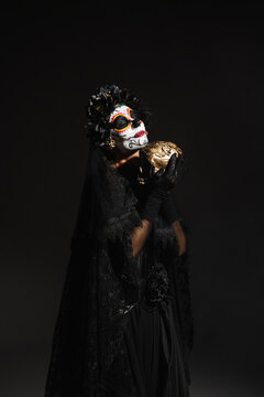 woman with sugar skull makeup wearing dark costume with veil and holding golden skull isolated on black.