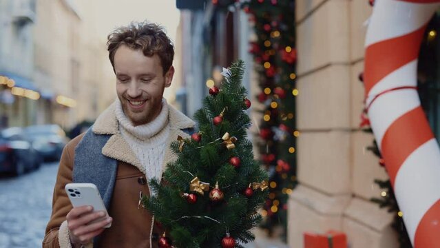 Smiled caucasian man going home from the christmas tree shop holding the little xmas tree using smartphone, swiping apps, texting with girlfriend. People and technology concept