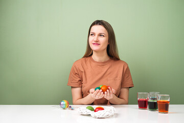A young beautiful woman in a brown t-shirt paints Easter eggs in different colors. Preparing Easter Eve at home. Green wall.