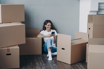 Happy girl choosing moving company for removal using phone, sitting with boxes. Mortgage, relocation