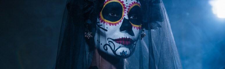 portrait of woman in halloween sugar skull makeup and veil on dark background with blue light, banner.