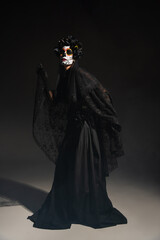 full length of woman in traditional day of dead costume and catrina makeup posing on black background.