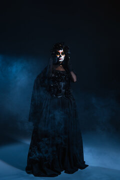 full length of woman in black costume and creepy halloween makeup posing with hand on waist on dark background with blue fog.
