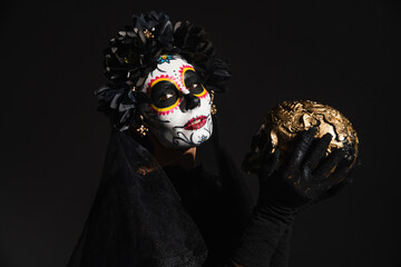 woman in sugar skull makeup and creepy dark costume holding golden skull isolated on black.