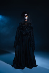 full length of woman in traditional santa muerte makeup and black dress standing in blue light on dark background.