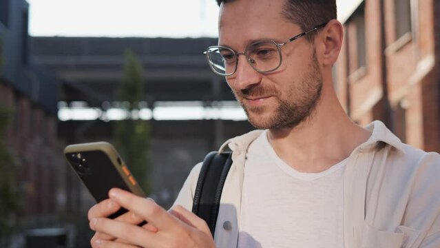 Close up portrait of business man male programmer in glasses standing outdoor in city and tapping on smartphone. Social media influencer using cellphone likes and comments posts. Blogger concept.
