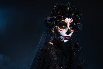 woman in traditional santa muerte makeup and black wreath holding hand near face on dark blue background.