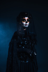 woman in scary halloween makeup and black costume with veil on dark blue background.