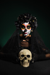 Woman in wreath with veil and catrina makeup looking at camera near skull on dark green background.