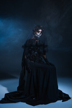 woman in halloween makeup and black witch dress sitting on dark smoky background with blue light.