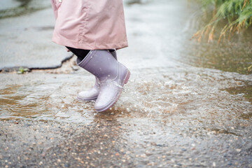 Little girl in pink waterproof raincoat, purple rubber boots funny jumps through puddles on street...