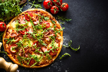 Pizza on black background. Traditional italian pizza with ham, cheese, tomatoes and arugula. Top view with copy space.
