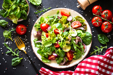 Salad leaves and vegetables in white plate at black table. Top view with ingredients.