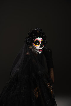 woman in spooky halloween makeup and black wreath with veil looking at camera on dark background.