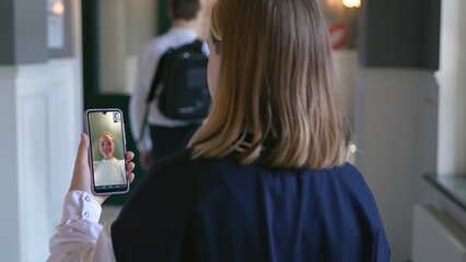 Back view of teen girl having video call with friend standing in school corridor