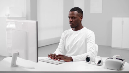 African-American man in white clothes typing on computer sitting at desk in white office