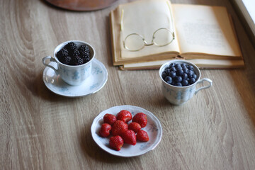Vintage tea set filled with blueberries, blackberries and starwberries, open book, reading glasses and decorative candles on the table. Selective focus.