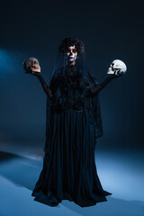 full length of woman in black costume and spooky halloween makeup holding skulls on dark background with blue light.