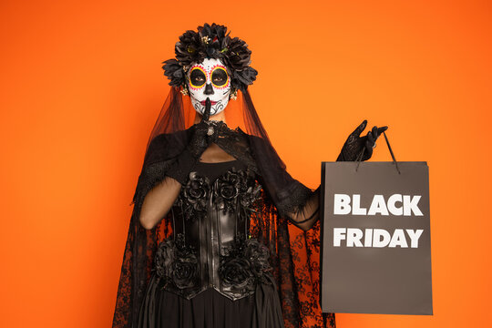 woman in sugar skull halloween makeup and costume showing hush and holding black friday shopping bag isolated on orange.