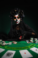KYIV, UKRAINE - SEPTEMBER 12, 2022: Woman in day of death costume throwing dice near playing cards and chips on dark green background.