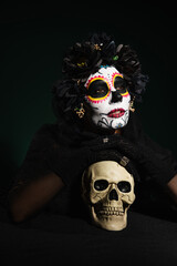 Woman in mexican day of dead makeup holding skull on dark green background.