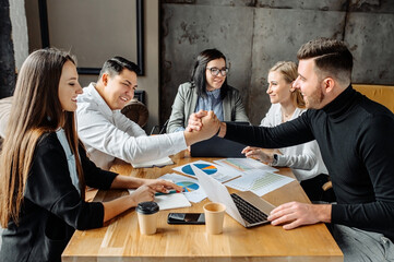 Successful deal. A group of young people are sitting in the office, two guys shake hands as a sign of a successful business transaction or sale. Everyone is smiling