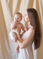 Young mother with long dark hair holding a cute little daughter in her arms in a nude fabric on a nude textile background. happy motherhood