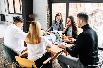 Two female colleagues discuss documentation at a table in a modern office at a meeting while their colleagues are working with laptops.