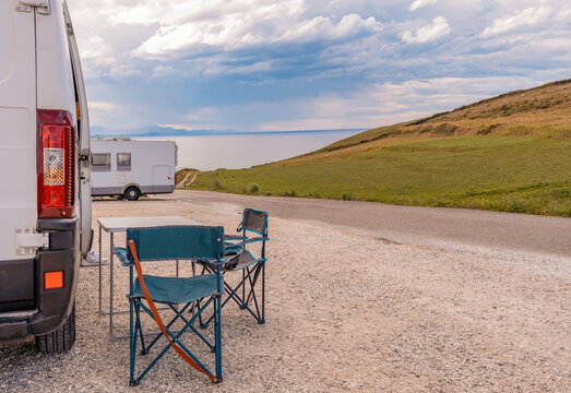 Vista de paisaje con mar y monta&ntilde;a, en camping con una camper con mesa y sillas y una caravana. Fotograf&iacute;a horizontal con copy space.