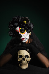 Woman in mexican halloween costume touching skull on dark green background.