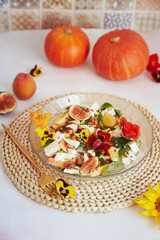 Fresh autumn vegetarian salad with edible flowers, figs, peach, greens and feta on ceramic tile background with fall pumpkin decorations.