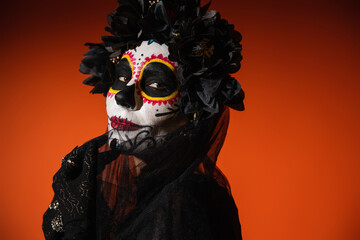 Portrait of woman with sugar skull makeup touching black veil on red background.