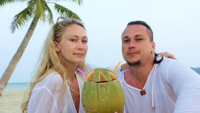 Cute loving couple facing each other sits in tropical beach under palm tree and drink coconat cocktail, laugh and having fun. Concept happy honeymoon relationship, happiness love