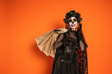 happy woman in mexican day of dead makeup and black costume standing with shopping bags on orange background.