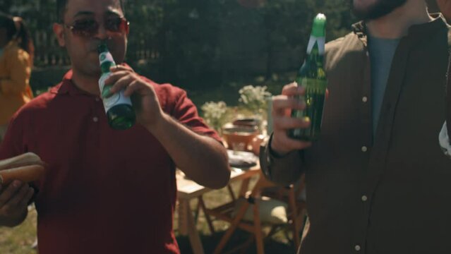 Multi-ethnic young men spending time together at outdoor bbq party clinking bottles with beer and enjoying drinking it
