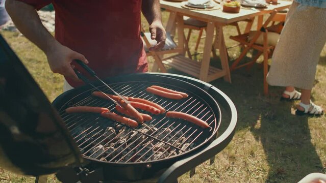 Unrecognizable man standing at barbecue grill cooking sausages for outdoor party on summer day