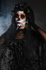 woman in sugar skull makeup and costume with black wreath and lace veil on dark background with smoke.