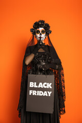 woman in creepy makeup and witch costume holding black friday shopping bag and showing hush sign isolated on orange.