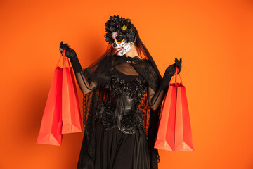 joyful woman in santa muerte makeup and black costume holding shopping bags isolated on orange.