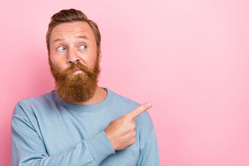 Portrait of positive interested impressed guy with red beard wear blue long sleeve directing empty space isolated on pink color background