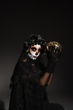 woman with traditional santa muerte makeup and costume with dark wreath holding golden skull on black background.