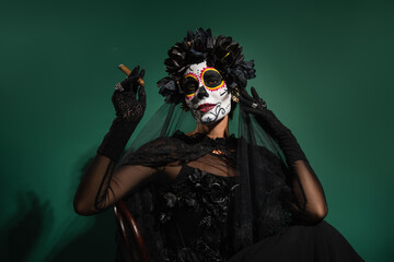 Woman with creepy santa muerte costume holding cigar and posing on green background.