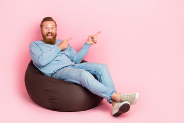 Full length portrait of astonished crazy man sit bag indicate fingers empty space isolated on pink color background