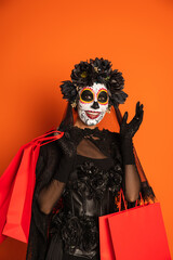 woman in sugar skull makeup and black halloween costume holding shopping bags and waving hand isolated on orange.