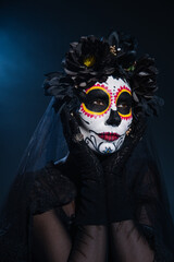 woman in black wreath holding hands near face with traditional santa muerte makeup on dark blue background.