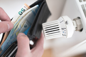 close-up view of person holding wallet with cash next to turned down thermostat on radiator, rising...