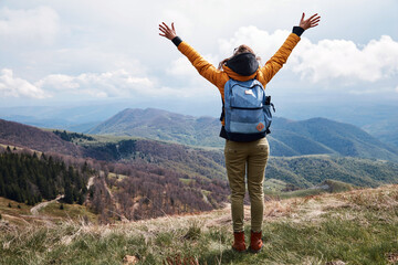 Young woman with rucksack and arms wide opened on the top of the hilly landscape.