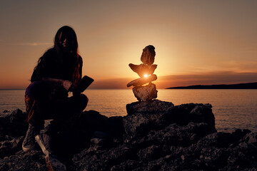 Silhouette of a woman balancing rocks and stones on the ocean sea coast at sunset sunrise time.