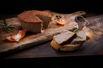 Round, homemade meat pate. Composition with a baked meat product on a rustic wooden chopping board, on black background.